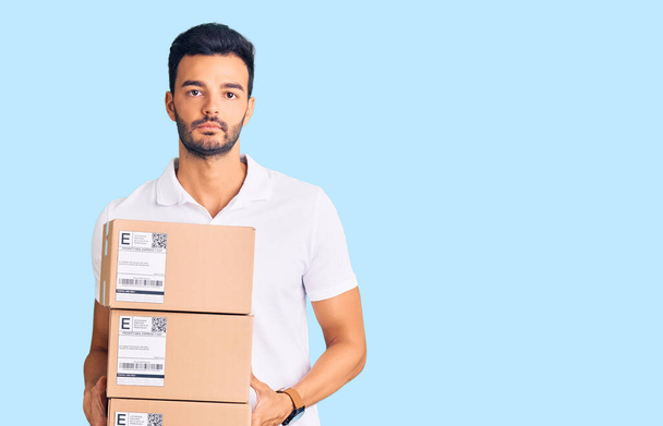 Young handsome hispanic man holding delivery package thinking attitude and sober expression looking self confident  - Photo, Image