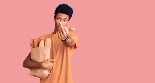Young african american man holding paper bag with bread showing middle finger, impolite and rude fuck off expression  - Fotografie, Obrázek