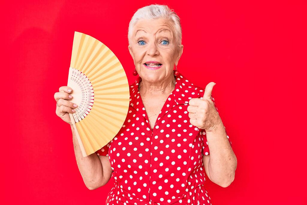 Senior mooie vrouw met blauwe ogen en grijs haar zwaaiende hand ventilator koeling lucht in de zomer glimlachen blij en positief, duim omhoog doen uitstekend en goedkeuring teken  - Foto, afbeelding