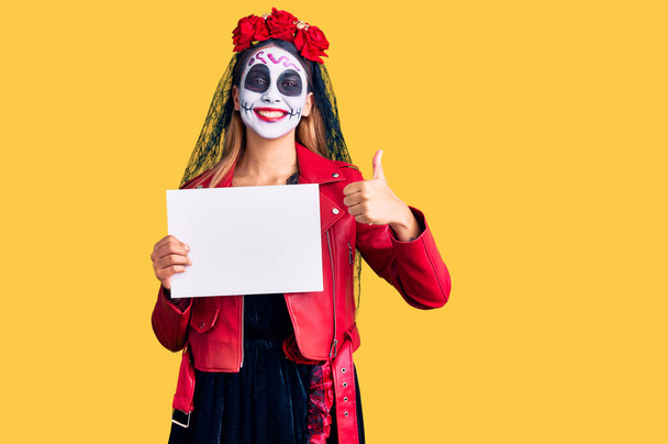 Mujer con el día del disfraz muerto sosteniendo banner vacío en blanco sonriendo feliz y positivo, pulgar hacia arriba haciendo excelente y signo de aprobación  - Foto, Imagen