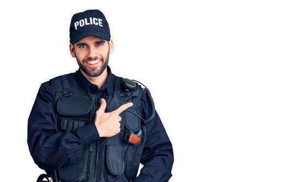 Jovem homem bonito com barba vestindo uniforme policial alegre com um sorriso de rosto apontando com a mão e o dedo para o lado com expressão feliz e natural no rosto  - Foto, Imagem