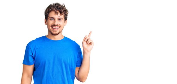 Young handsome man with curly hair wearing casual clothes with a big smile on face, pointing with hand finger to the side looking at the camera.  - Photo, Image
