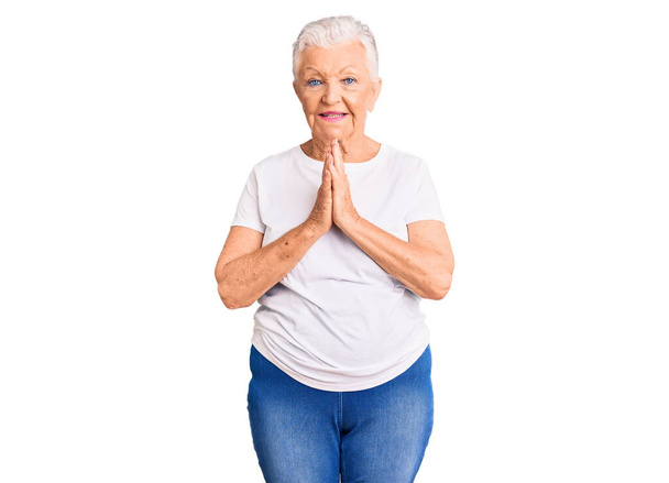 Starsza piękna kobieta z niebieskimi oczami i siwymi włosami ubrana w luźną białą koszulę modląca się razem z dłońmi prosząc o przebaczenie uśmiechając się pewnie.  - Zdjęcie, obraz