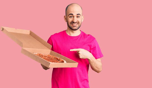 Joven hombre guapo sosteniendo entrega pizza caja de cartón sonriendo feliz señalando con la mano y el dedo  - Foto, Imagen