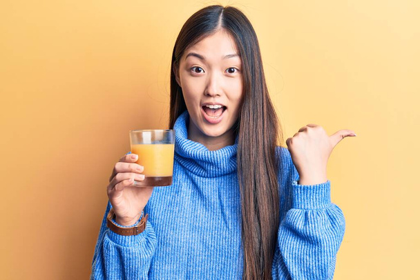Jovem mulher chinesa bonita bebendo copo de suco de laranja apontando polegar até o lado sorrindo feliz com a boca aberta
  - Foto, Imagem