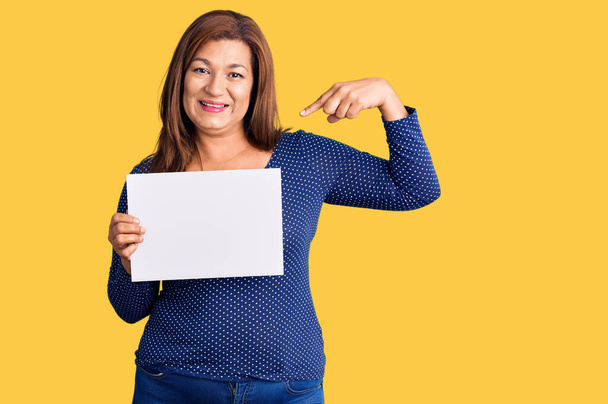 Латиноамериканка средних лет держит бумажный баннер с пустым местом, указывая пальцем на себя улыбающейся счастливой и гордой  - Фото, изображение