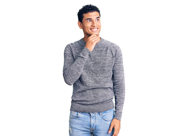 Hispanic knappe jongeman draagt casual trui met de hand op kin denken over vraag, penseelachtige uitdrukking. glimlachend met een bedachtzaam gezicht. twijfelconcept.  - Foto, afbeelding