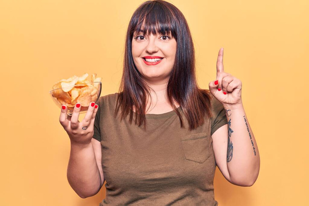 Young plus size woman holding potato chip surprised with an idea or question pointing finger with happy face, number one  - Photo, Image