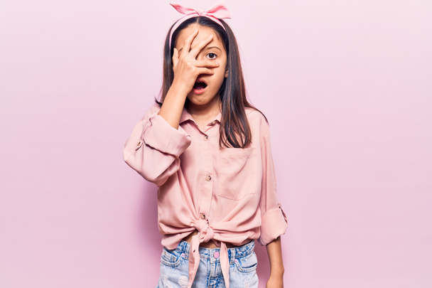 Beautiful child girl wearing casual clothes peeking in shock covering face and eyes with hand, looking through fingers with embarrassed expression.  - Foto, Bild