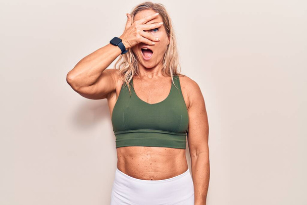 Middle age caucasian blonde woman wearing sportswear peeking in shock covering face and eyes with hand, looking through fingers with embarrassed expression.  - Φωτογραφία, εικόνα