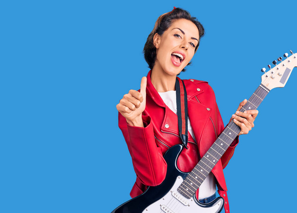 Νεαρή όμορφη γυναίκα παίζει ηλεκτρική κιθάρα χαμογελώντας χαρούμενος και θετικός, αντίχειρας επάνω κάνει εξαιρετική και την έγκριση σημάδι  - Φωτογραφία, εικόνα