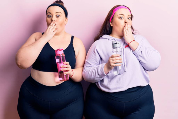 Young plus size twins wearing sportswear drinking bottle of water covering mouth with hand, shocked and afraid for mistake. surprised expression  - Foto, afbeelding