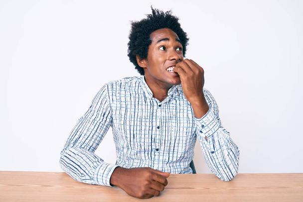 Schöner afrikanisch-amerikanischer Mann mit Afrohaaren in lässiger Kleidung, der auf dem Tisch sitzt und gestresst und nervös aussieht, die Hände auf den Mund beißenden Nägeln. Angstproblem.  - Foto, Bild