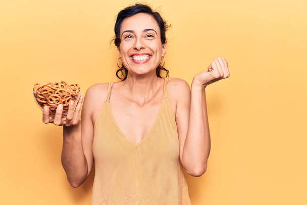 Young beautiful hispanic woman holding pretzels screaming proud, celebrating victory and success very excited with raised arm  - Photo, Image
