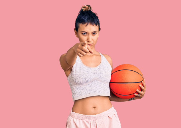若いです女性保持バスケットボールボールポインティングとともに指でカメラとあなたに、自信を持ってジェスチャーを真剣に見て  - 写真・画像