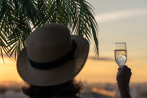 Back view of lonely beautiful girl with glass of champagn  in a straw hat against the background of the sea in branches of palm trees. Sunset beach. Summer and freedom concept. - Fotó, kép