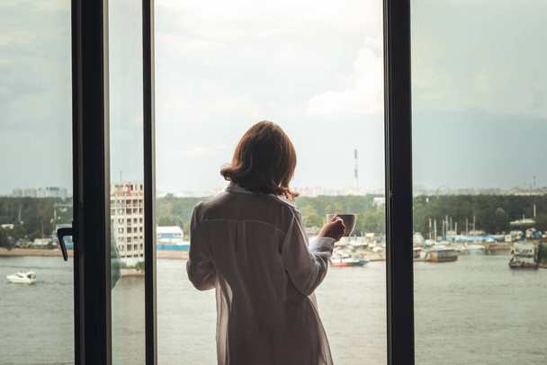Back view of beautiful young woman with cup of coffee in a transparent nightgown standing and enjoying near open panoramic window of  apartments.  - Φωτογραφία, εικόνα