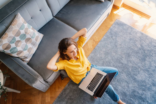 Beautiful young girl woman in yellow shirt works on laptop at home in living room sitting on floor, top view image - Фото, изображение
