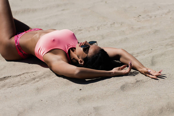 Trendy female model in pink top and panties lying on sand and looking away while relaxing on beach on sunny summer day - 写真・画像