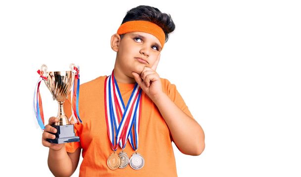 Pieni poika, jolla on mestaripalkinto ja jolla on mitalit vakavat kasvot ajattelevat kysymystä käsi leualla, harkitsee hämmentävää ideaa.  - Valokuva, kuva