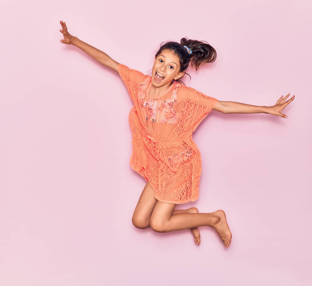 Adorable hispanic child girl on vacation wearing summer dress smiling happy. Jumping with smile on face over isolated pink background - Фото, изображение