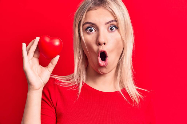 Young beautiful blonde woman holding plastic heart standing over isolated red background scared and amazed with open mouth for surprise, disbelief face - Φωτογραφία, εικόνα
