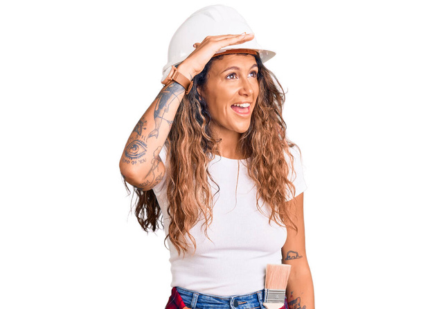 Young hispanic woman with tattoo wearing hardhat and builder clothes very happy and smiling looking far away with hand over head. searching concept.  - Photo, Image