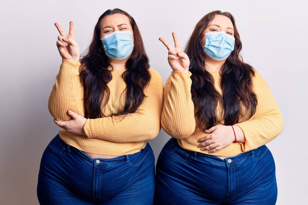 Young plus size twins wearing medical mask smiling with happy face winking at the camera doing victory sign. number two.  - Photo, Image