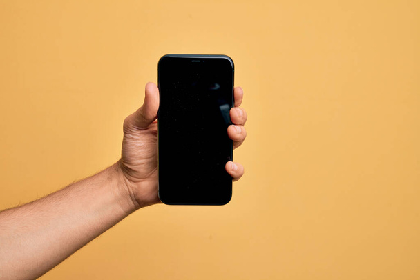 Hand of kaukázusi fiatal férfi holding smartphone bemutató képernyő felett elszigetelt sárga háttér - Fotó, kép