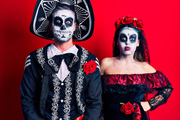 Young couple wearing mexican day of the dead costume over red puffing cheeks with funny face. mouth inflated with air, crazy expression.  - Photo, Image