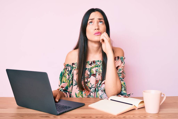Young caucasian woman sitting at the desk studying using laptop and book drinking coffee serious face thinking about question with hand on chin, thoughtful about confusing idea  - Foto, imagen