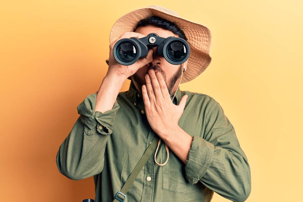 Young hispanic man wearing explorer hat holding binoculars covering mouth with hand, shocked and afraid for mistake. surprised expression  - Photo, Image