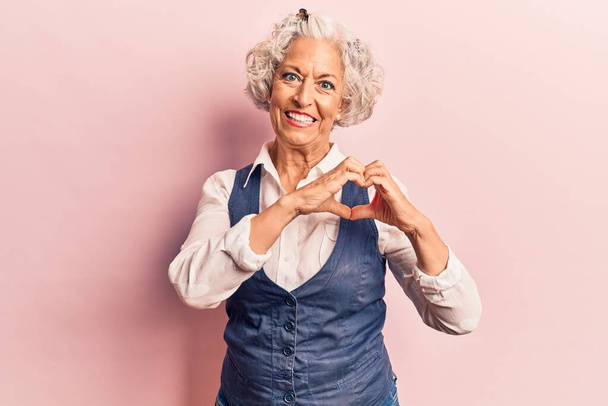 Eine ältere grauhaarige Frau in lässiger Kleidung, die verliebt lächelt und mit den Händen Herzsymbole in Form bringt. Romantisches Konzept.  - Foto, Bild