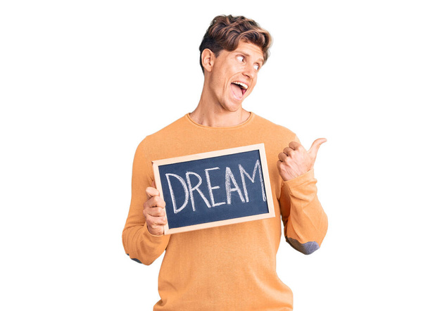 Young handsome man holding blackboard with dream word pointing thumb up to the side smiling happy with open mouth  - Foto, imagen