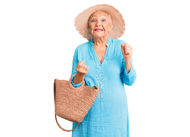 Старшая красивая женщина с голубыми глазами и седыми волосами в модном платье и шляпе держа летнюю плетеную сумочку гордо крича, празднуя победу и успех очень взволнован с поднятыми руками  - Фото, изображение