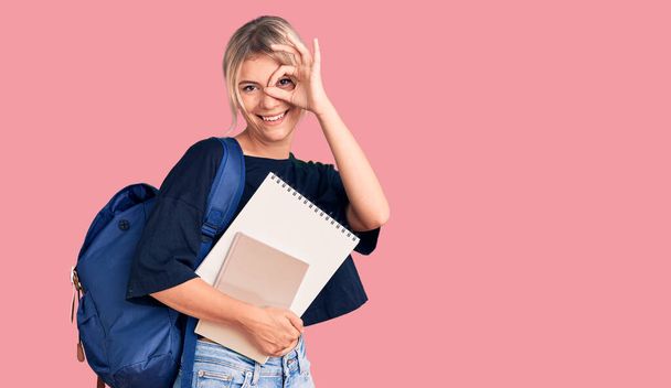 Fiatal gyönyörű szőke nő visel diák hátizsák gazdaság notebook mosolyogva boldog csinál rendben jel kézzel a szemen keresztül néz az ujjakon  - Fotó, kép