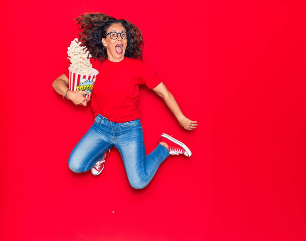 Middelbare leeftijd mooie Latijns-Amerikaanse vrouw verrast met open mond dragen van een bril. emmer popcorn die over een geïsoleerde rode achtergrond springt - Foto, afbeelding