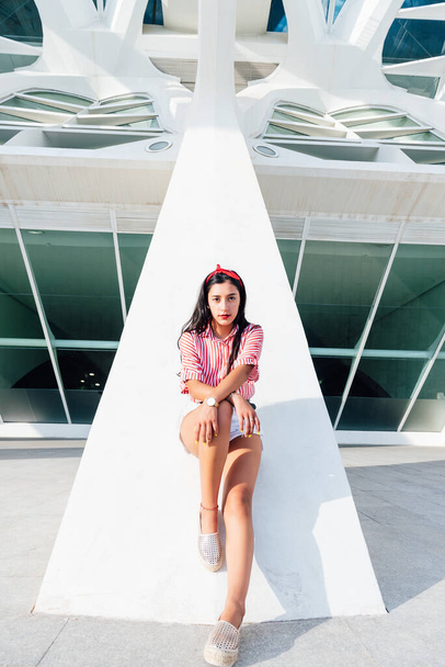 Chica latina joven con el pelo negro y cinta roja en el pelo, de pie mirando a la cámara en un día soleado con fondo blanco, vistiendo camisa de rayas rojas. espacio de copia - Foto, Imagen