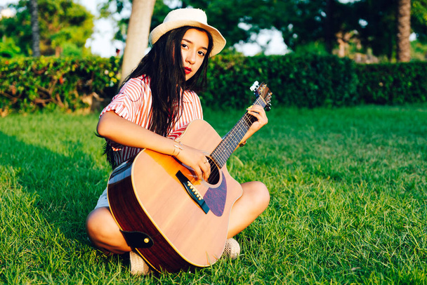 Молодая длинноволосая латиноамериканка в красно-белой полосатой рубашке и шляпе, сидящая на садовом газоне и играющая на акустической гитаре. копировальное пространство - Фото, изображение
