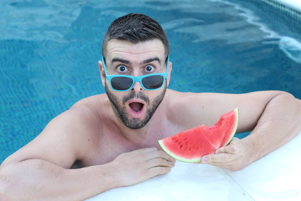 Nahaufnahme Porträt eines hübschen jungen Mannes im Swimmingpool mit einer Scheibe Wassermelone, die in die Kamera blickt - Foto, Bild