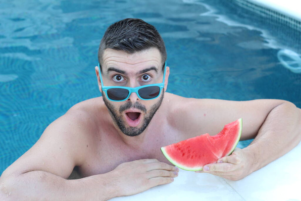 close-up portrait of handsome young man in swimming pool with slice of watermelon looking at camera - Photo, Image