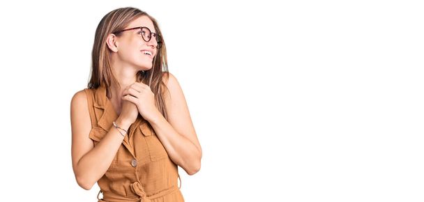 Young beautiful blonde woman wearing casual clothes and glasses laughing nervous and excited with hands on chin looking to the side  - Foto, afbeelding