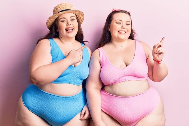 Young plus size twins wearing bikini smiling and looking at the camera pointing with two hands and fingers to the side.  - Photo, Image