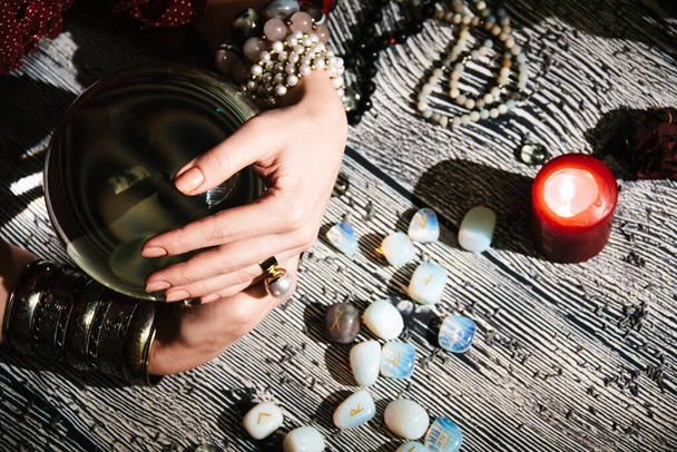Fortuneteller's hands on a glass orb. Prediction of the future. Mystic interior. Occult symbols, runes stones, rosaries, candles - Photo, image