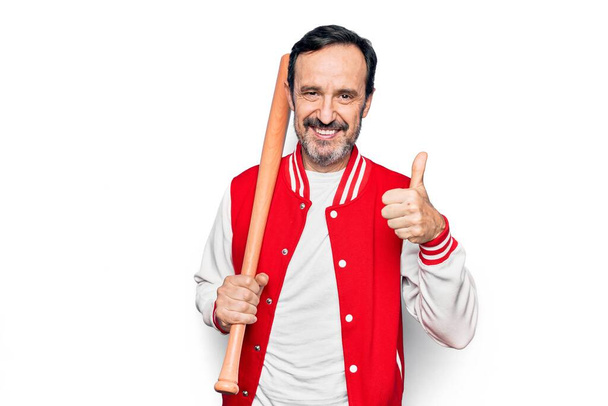 Középkorú jóképű sportos férfi visel kabát baseballt használ fa denevér mosolygós boldog és pozitív, hüvelykujj fel csinál kiváló és jóváhagyási jel - Fotó, kép