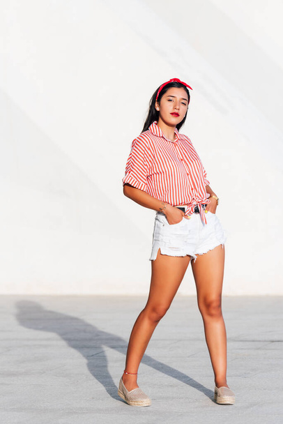 Menina latina jovem com cabelo preto e fita vermelha em seu cabelo, de pé olhando para a câmera em um dia ensolarado com um fundo branco, vestindo uma camisa listrada vermelha e branca e calças curtas. espaço de cópia - Foto, Imagem