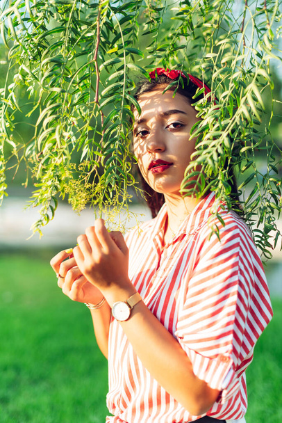Mladá černosrstá latinskoamerická dívka s červenou stuhou ve vlasech a červenobílou pruhovanou košilí, s hlavou na listech stromu, dívá se do kamery - Fotografie, Obrázek