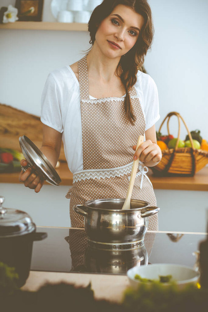 Young brunette woman cooking soup in kitchen. Housewife holding wooden spoon in her hand. Food and health concept - Photo, Image