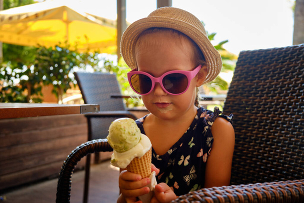 Little girl dressed in straw hat and pink sunglasses eats ice cream at cafe. - 写真・画像