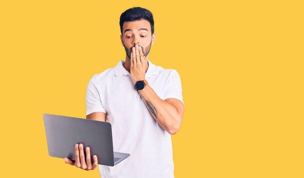 Junge gut aussehende hispanische Mann arbeitet mit Computer-Laptop bedeckt Mund mit der Hand, schockiert und Angst vor Fehler. Überraschte Miene  - Foto, Bild